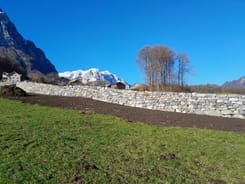 Trockenmauer in offener Landschaft im Kanton Glarus mit Wiese, Häusern, kahlen Bäumen und schneebedeckten Bergen.