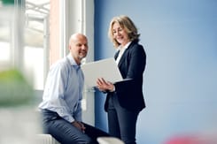 Eine Frau zeigt einem sitzenden Mann Inhalte auf einem Laptop während eines persönlichen Gesprächs im Büro.