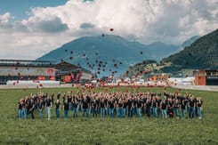 Gruppenfoto von Mitarbeitenden auf einer Wiese vor Bergkulisse beim gemeinsamen Werfen roter GLKB-Kappen.