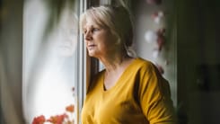 Ältere Frau im senfgelben Pullover blickt nachdenklich aus dem Fenster in ihrem Zuhause.