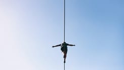 Mann balanciert auf einem Hochseil in grosser Hoehe vor blauem Himmel mit ausgestreckten Armen.