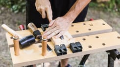 Handwerker bearbeitet Holzstueck mit Stechbeitel auf Werkbank, umgeben von Werkzeugen im Freien.