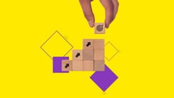 Hand setzt letzten Holzbaustein mit Zielsymbol auf gestapelte Würfel mit Pfeilen auf gelbem Hintergrund.