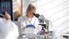 Wissenschaftlerin im Labor analysiert Proben mit Mikroskop, umgeben von Reagenzglaesern und Laptop.