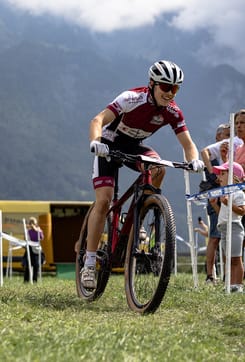 Mountainbiker fährt bei einem Wettkampf über eine Wiese vor Bergkulisse und Zuschauern im Hintergrund