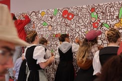 Ehrendamen in Trachten und Kinder bemalen gemeinsam eine GLKB-Wand am Promostand der GLKB.