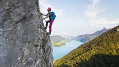 Kletterer mit Helm und Ausruestung erklimmt steile Felswand mit Blick auf See und Berglandschaft.