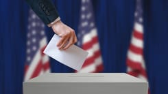 Hand steckt Wahlzettel in eine Urne vor unscharfen US-Flaggen als Symbolbild fuer demokratische Wahl.