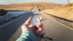 Hand haelt Kompass in den Vordergrund, waehrend eine leere Landstrasse in die Ferne fuehrt.