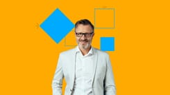 Älterer Mann mit Brille und grauem Bart in hellem Anzug vor orangefarbenem Hintergrund mit blauen Formen.