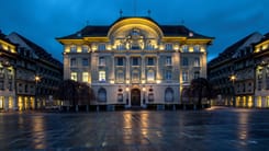 Beleuchtetes historisches Bankgebaeude bei Abenddaemmerung auf leerem Platz inmitten einer Altstadt.