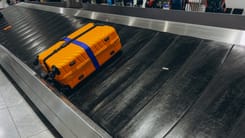 Ein orangefarbener Reisekoffer mit blauem Gurt liegt auf einem Gepäckband am Flughafen.