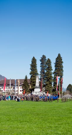 Menschenmenge bei der Näfelser Fahrt auf grüner Wiese vor Denkmal, Fahnen und hohen Bäumen unter blauem Himmel.