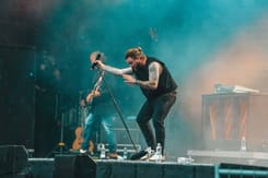 Tätowierter Sänger beugt sich mit Mikrofon im Regen bei energiegeladener Bühnenperformance.