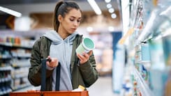 Frau im Supermarkt betrachtet aufmerksam ein Produkt in der Hand mit Einkaufskorb im Vordergrund.