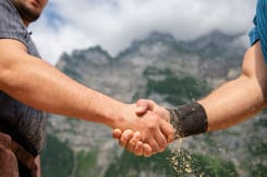 Nahaufnahme eines traditionellen Handschlags zweier Schwinger vor Bergkulisse mit Sägemehl in der Luft.