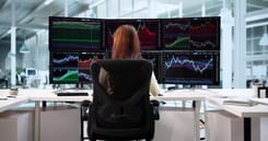 Frau beobachtet mehrere Monitore mit Börsenkursen und Charts in einem modernen Trading- oder Analysebüro.