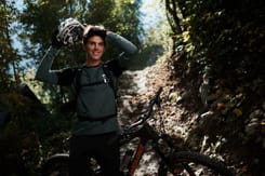 Mountainbiker steht im Wald auf einem schmalen Trail und hält seinen Helm in der Hand.