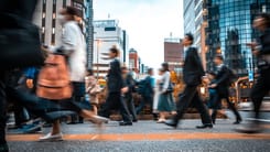 Menschenmengen ueberqueren eine Strasse in geschäftiger Grossstadt mit Hochhaeusern im Hintergrund.
