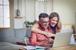 Lächelndes Paar nutzt gemeinsam ein Notebook zu Hause für Online-Banking oder digitale Finanzplanung.