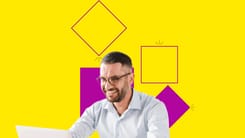 Mann mit Brille und Bart arbeitet lächelnd am Laptop vor gelbem Hintergrund mit pinken Formen.