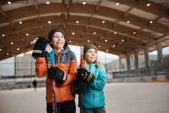Zwei Kinder in Winterkleidung mit Eishockeyschlägern auf der überdachten Eislaufbahn in der GLKB Arena.