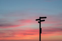 Ein Wegweiser der in der Natur steht. Im Hintergrund sieht man den Sonnenuntergang. 