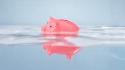 Rosa Sparschwein treibt symbolisch auf Wasseroberflaeche mit Spiegelung im ruhigen Blau.