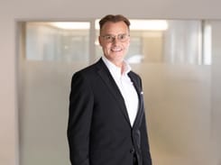Portrait von Roland Wickart im Anzug vor einem modernen, lichtdurchfluteten Bürohintergrund.