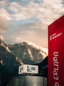 Schriftzug «Mitenand erläbä» vor imposanter Glarner Bergkulisse bei klarem Himmel.