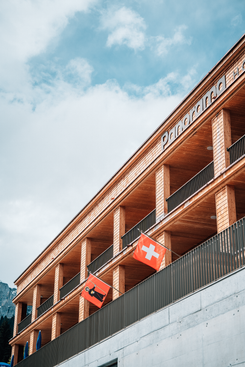 Gebäude mit Holzfassade und Schweizer Fahnen vor Bergkulisse, aufgenommen während eines Aufenthalts oder Firmenevents.