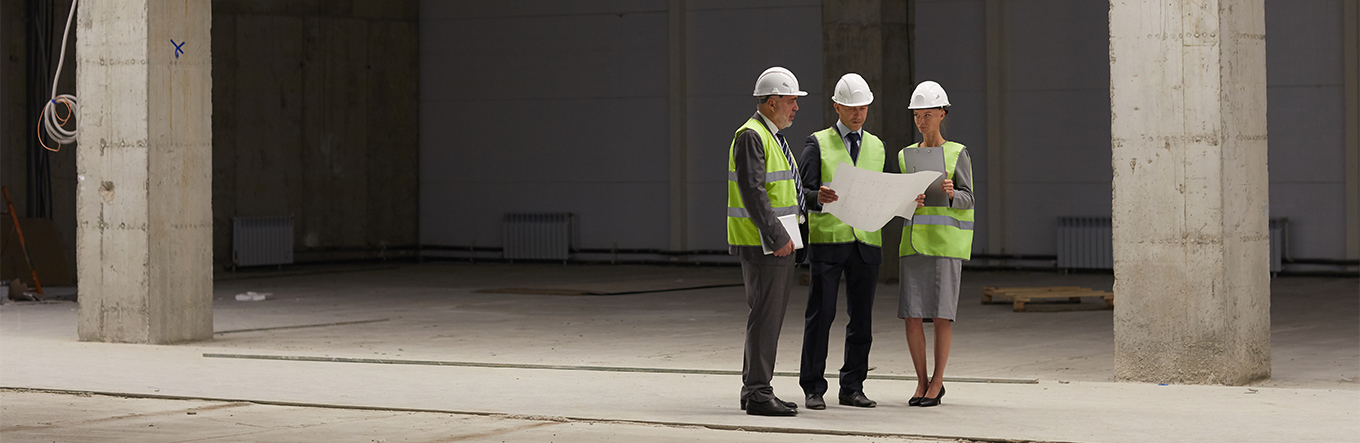 Zwei Geschäftsmänner in Anzug und eine Geschäftsfrau elegant gekleidet unterhalten sich auf einer Baustelle über einen Plan in Papierform und tragen sowohl Sicherheitshelme als auch Sicherheitswesten 